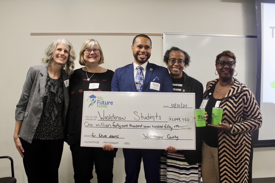 Five people stand smiling while holding a big check written out to students and their futures.