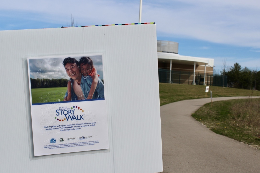 Photo of a sign staked into the ground with information written in text and Braille about the Story Walk along a walking path